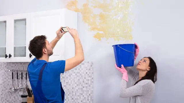 Couple photographing a water stain on a wall while catching a drip in a blue bucket for an insurance claim