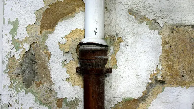 Corroded pipe running through a wall with peeling, stained plaster from a hidden leak